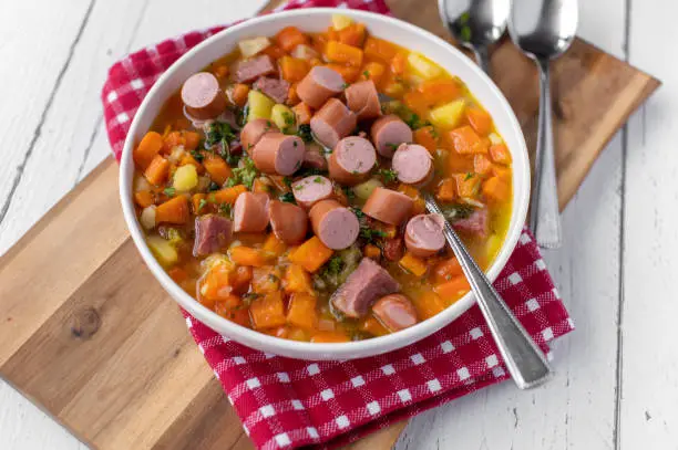 Delicious homemade carrot soup or stew with pork meat and vienna sausage. Served hot and ready to eat on deep plate with spoon isolated on light background. Closeup