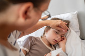 Sick child sleeping at home in bed with flu, cold, coronavirus, bronchitis or pneumonia, virus or infection and his mother touching his forehead checking temperature