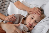 Sick child sleeping at home in bed with flue, cold, covid, bronchitis or pneumonia, virus or infection and his mother's hand touching his forehead checking temperature