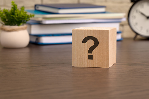 question mark is written on wooden cube lying on the table, in the background there is a stack of textbooks, an alarm clock and a potted plant. cube close up