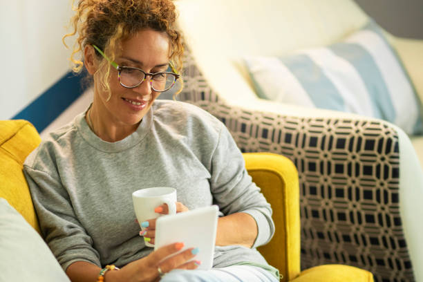 istock Pretty woman enjoy relax at home reading a book on tabler reader and drinking a tea or coffe sitting on a yellow sofa in living room - modern online library store and female people using device gadget to buy and read 1920410019