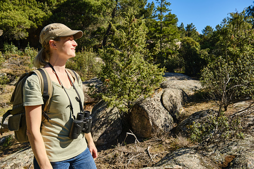 Side view of young female traveler with backpack and binoculars standing in green forest on sunny day while looking away