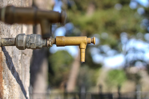 Park faucet without water, with drop on the tip, unfocused park background, side view. Water shortage concept