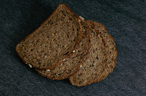 Four slices of rye bread with fiber and sunflower seeds lie on a black stone table with a blurred background, flat lay close-up. The concept of baking bread.