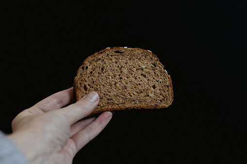 The hand of a young caucasian girl holds one piece of rye bread with sunflower seeds on a black background, close-up side view. The concept of baking bread.