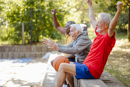 Photo of vital seniors in sports outfits on a basketball court. They are watching basketball game and supporting thir team