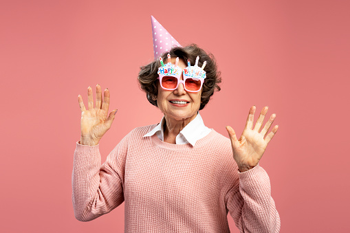 Smiling beautiful senior 70 years old woman wearing funny wearing eyeglasses hands up isolated on pink background. Celebration birthday concept