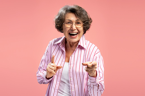 Smiling senior 70 years old woman wearing eyeglasses and stylish casual clothes pointing fingers isolated on pink background. Having fun concept