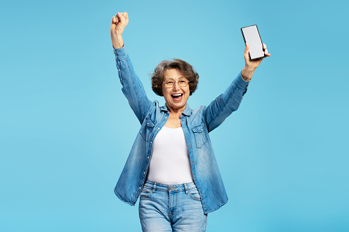 Overjoyed senior woman holding mobile phone with mockup, win something celebration success isolated on blue background