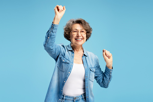 Attractive smiling senior, happy modern grandmother woman holding hands up dancing celebration success isolated on blue background. Good news, positive lifestyle concept