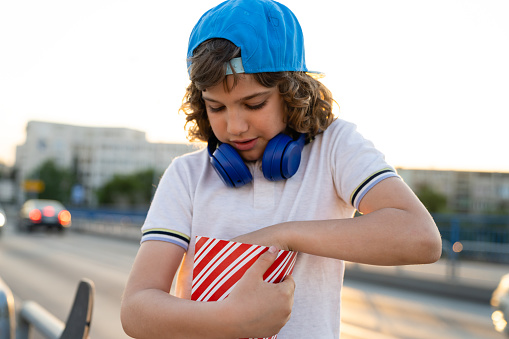 A boy with a blue cap and blue headphones around his neck is eating popcorn outside, on a bridge. Skater boy eating popcorn outside. Active children are happy children. Active teenage boy.