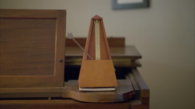 An antique vintage wooden metronome set to 42 beats per minute swings slowly back and forth to mark time on top of an old brown piano