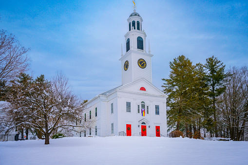 A tall white church with a clock tower, rainbow flag, and red doors on the snow-covered hilltop