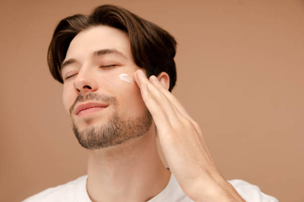 istock Close up of smiling man applying face cream, posing in studio. Isolated on beige background. Skin care, cosmetic, spa treatment concept 1920881000