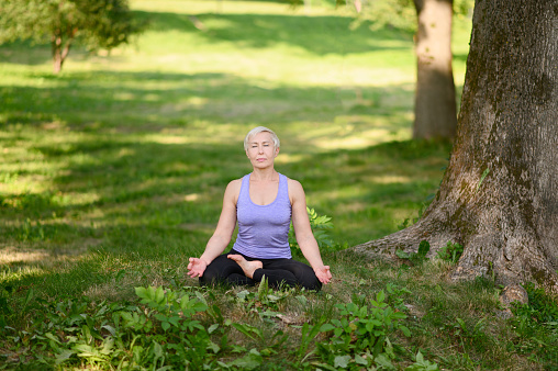 Athletic middle-aged woman doing yoga outdoors in the city park in the lotus position with her eyes closed. The concept of meditation, active lifestyle.