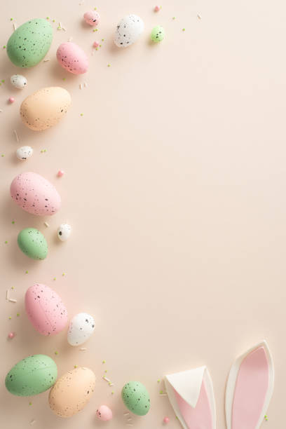 istock Festive Egg Ensemble: Top view vertical image featuring festive Easter eggs, bunny ears, and confectionery sprinkles on a pastel beige surfaceâperfect for text or promotional messages 1920924525