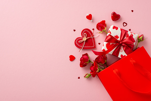 Romantic surprise awaits! Top view shot of paper bag with themed giftbox, adorned with love-patterned paper. Red roses, heart-shaped confetti on pastel pink backdrop, leaving space for sweet message