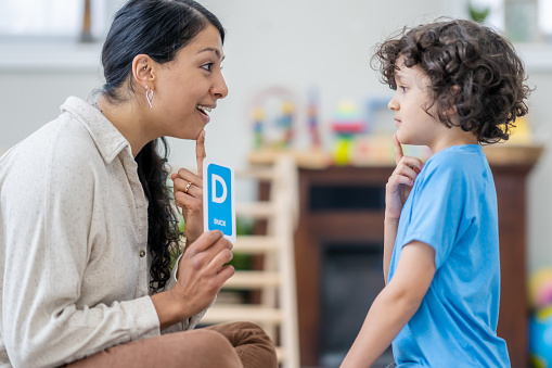 A female speech therapist works with a young boy as they practice their sounds and letter enunciation.  The Therapist is dressed casually and is holding up alphabetical cue cards as they work together.