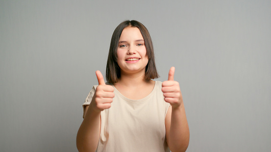 Image of confident latin teenager woman showing thumb up in approval, recommending, like smth good, standing over grey background.