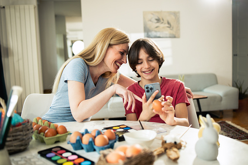 Portrait of a happy woman and her son painting Easter eggs at home and taking photos with a smartphone