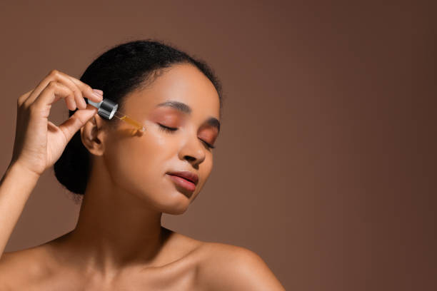 istock Beautiful woman applying serum onto her face on brown background. Space for text 1921020501