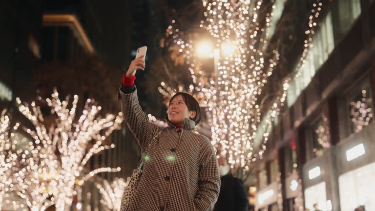 Woman taking photos and videos of Christmas lights with her mobile smart phone in city at night