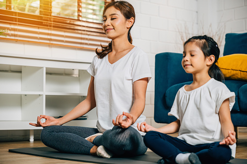 A mother teaches her little daughter yoga and meditation focusing on lotus pose and mudra gesture. This family's togetherness and balance create a peaceful and joyful moment at home.