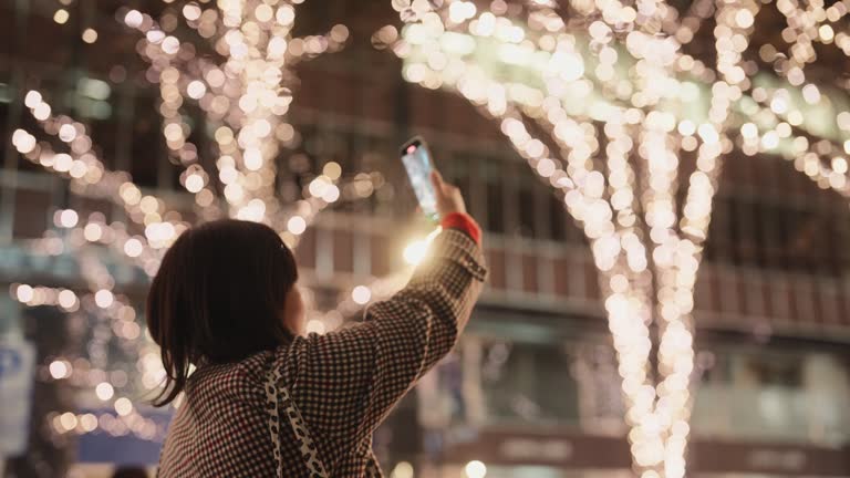 Woman taking photos and videos of Christmas lights with her mobile smart phone in city at night