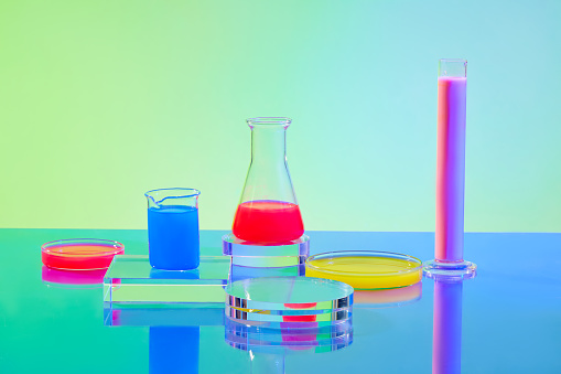 Red, pink, yellow and blue liquids inside glassware and transparent platforms on a green-blue gradient background. Lab space for advertising.