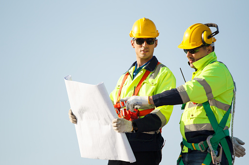 Senior professional engineers in full protection suit discuss on a blueprint paper on factory or building rooftop. Technicians inspecting the installation on rooftop.