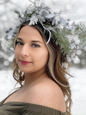 Headshot of beautiful young adult woman, outdoors in winter, wearing a head crown made out of natural elements, blending into the winter surroundings with Mother Nature vibes. She is looking at the camera, smiling.