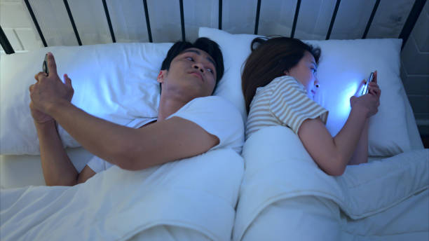 young couple sleeping on the bed in the bedroom at night time with their backs to each other and playing on their phones, ignoring each other. - sleeping lying on back couple bed photos et images de collection