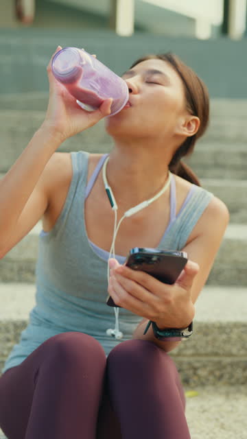 Young Asian female athlete runner drinking protein shake and using smartphone after exercise workout in urban park. Diet and healthy food workout exercise. Vertical Screen.