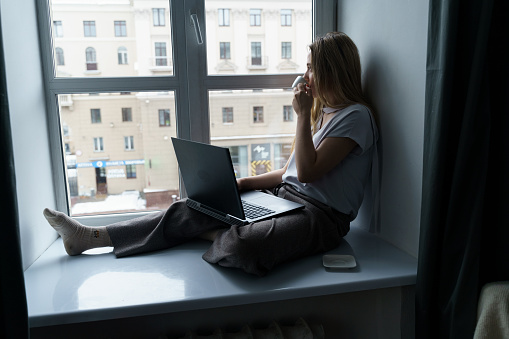A young woman sits on a windowsill, drinks coffee, works at a laptop, looks out the window. Business and education concept.