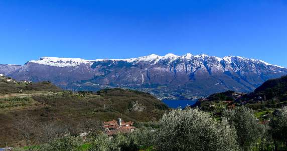 View from Tremosine to the snow-covered mountain range of the Monde Baldo mountains. In the foreground, olive trees and the village of Priezzo, a district of Tremosine. You can also see a small section of Lake Garda.