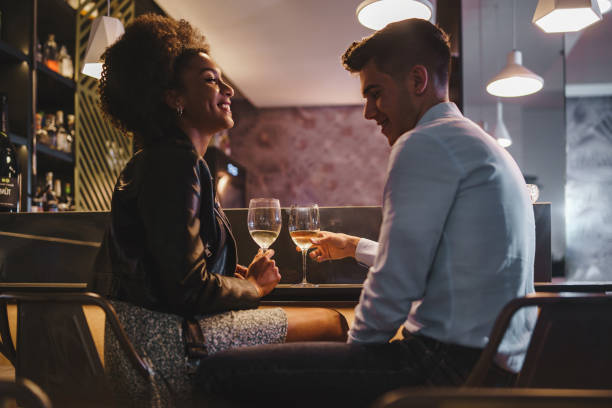 Romantic couple on a date talking holding a glass of white wine sitting at the bar counter in the night Romantic couple on a date talking holding a glass of white wine sitting at the bar counter in the night - food and drink, people, lifestyle concept black man white woman couple stock pictures, royalty-free photos & images