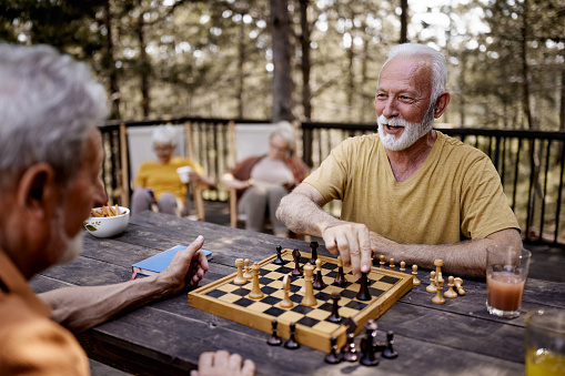 Happy mature man talking to his friend while playing chess on a terrace. Women are in the background.