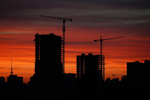 Sunset against the backdrop of a large construction site with several working cranes and unfinished high-rise buildings. Sunset with orange and red sky in cityscape with silhouettes of buildings