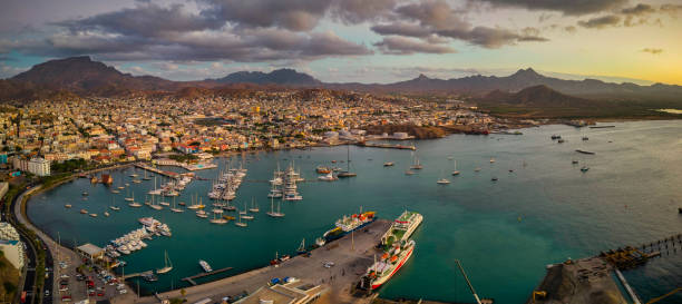 Spectacular Sunset Over Mindelo: A Panoramic Aerial View Panoramic aerial view of Mindelo city at sunset, with the marina and boats in the foreground, surrounded by the vibrant cityscape and mountains in the background under a warm, glowing sky west africa coast photos stock pictures, royalty-free photos & images