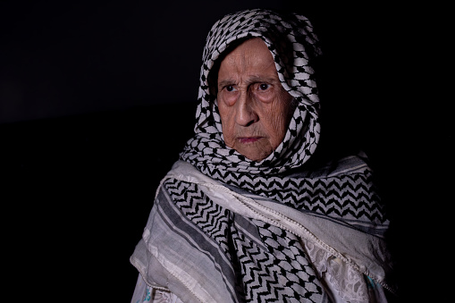 portrait of an old lady in dark background wearing white palestinian keffiyeh with smile on her face