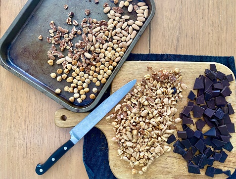 Horizontal flat lay of ingredients of chopped roasted nuts and chocolate ready to make Italian panforte dessert with cutting board knife and baking tray in domestic kitchen.