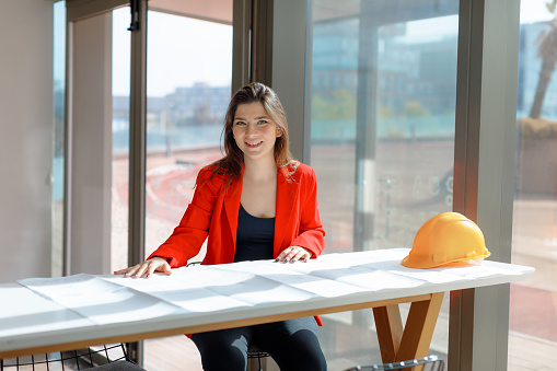 Portrait of the Happy and Successful beauty and satisfaction of a young and accomplished businesswoman. Dressed in a fashionable black shirt and a vibrant red blazer, she exudes confidence and professionalism. With her eyeglasses on, she smiles at the camera while working on her laptop and reviewing paperwork, surrounded by the creative atmosphere of a modern office. The sunlight streaming through the window adds a touch of brightness to the scene, reflecting her expertise in the fields of engineering, architecture, and business. Fashionable, black shirt, red blazer, eyeglasses, desk, laptop, paperwork, blueprint, modern office, sunlight, creative, engineering, architecture, business, agency, confidence, professionalism, success concept