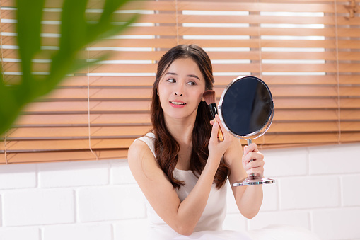 Butiful asian woman wearing pajamas applying makeup while sitting on bed with a mirror.