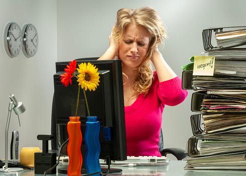 Young woman in office working late, with stress behind her monitor. Hands on head.