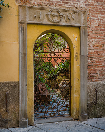 A metal garden gate with terracotta pots, brick wall and carved surround, in a quiet street in Lucca, Italy.