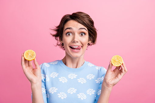 Photo of cheerful pretty girl tongue lick teeth arms hold two juicy lemon halves isolated on pink color background.