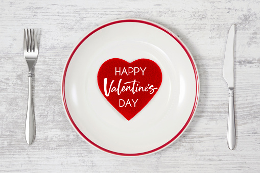 Felt red heart in white plate with fork and table knife on white wooden background for Valentine’s Day concept