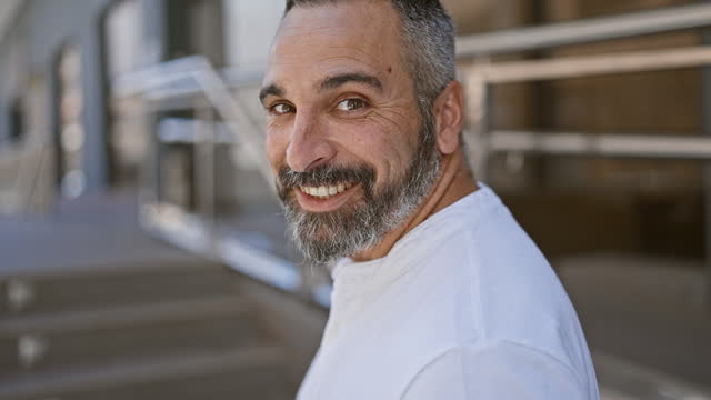 A smiling bearded mature man in a white shirt stands on an urban street with a blurred building backdrop.