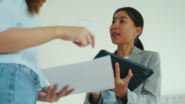 Close-up of Asian female real estate agent explaining and showing living room in new house to tenants for rent or buy. Landlord selling real estate to happiness couple customer.