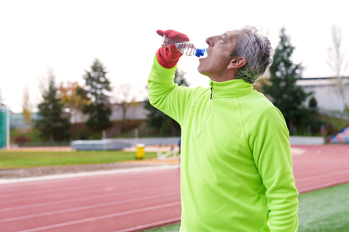 Elderly man in a bright lime sweatshirt hydrates with a water bottle during his workout on an outdoor running track